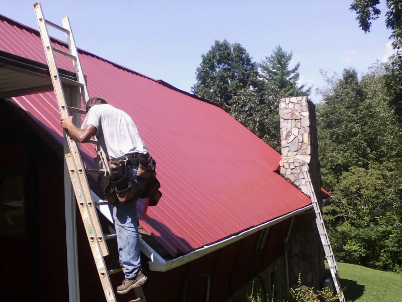 Roofer on a ladder beside a red metal roof and stone chimney — Soffit & Fascia Repair by Roofing Brothers in Washoe Valley